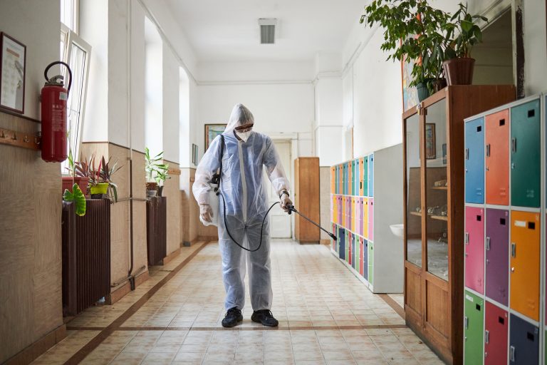 Man with protective suit with surgical gloves, n95 face mask and protective eyewear, spraying in a school hallway